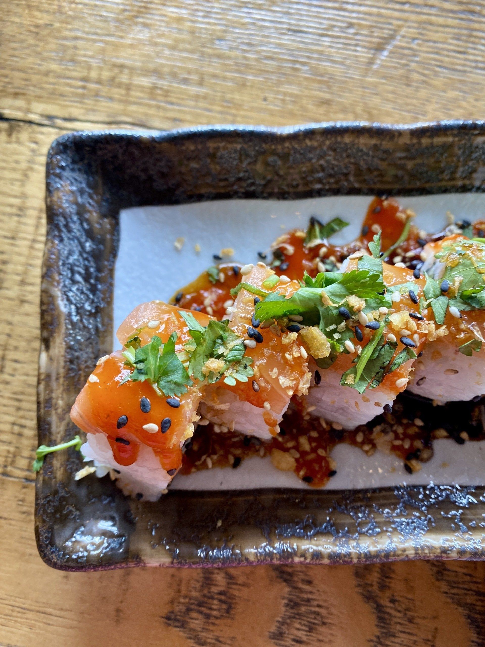 Sushi rolls with salmon, sauce, sesame seeds, and cilantro on a rectangular plate.