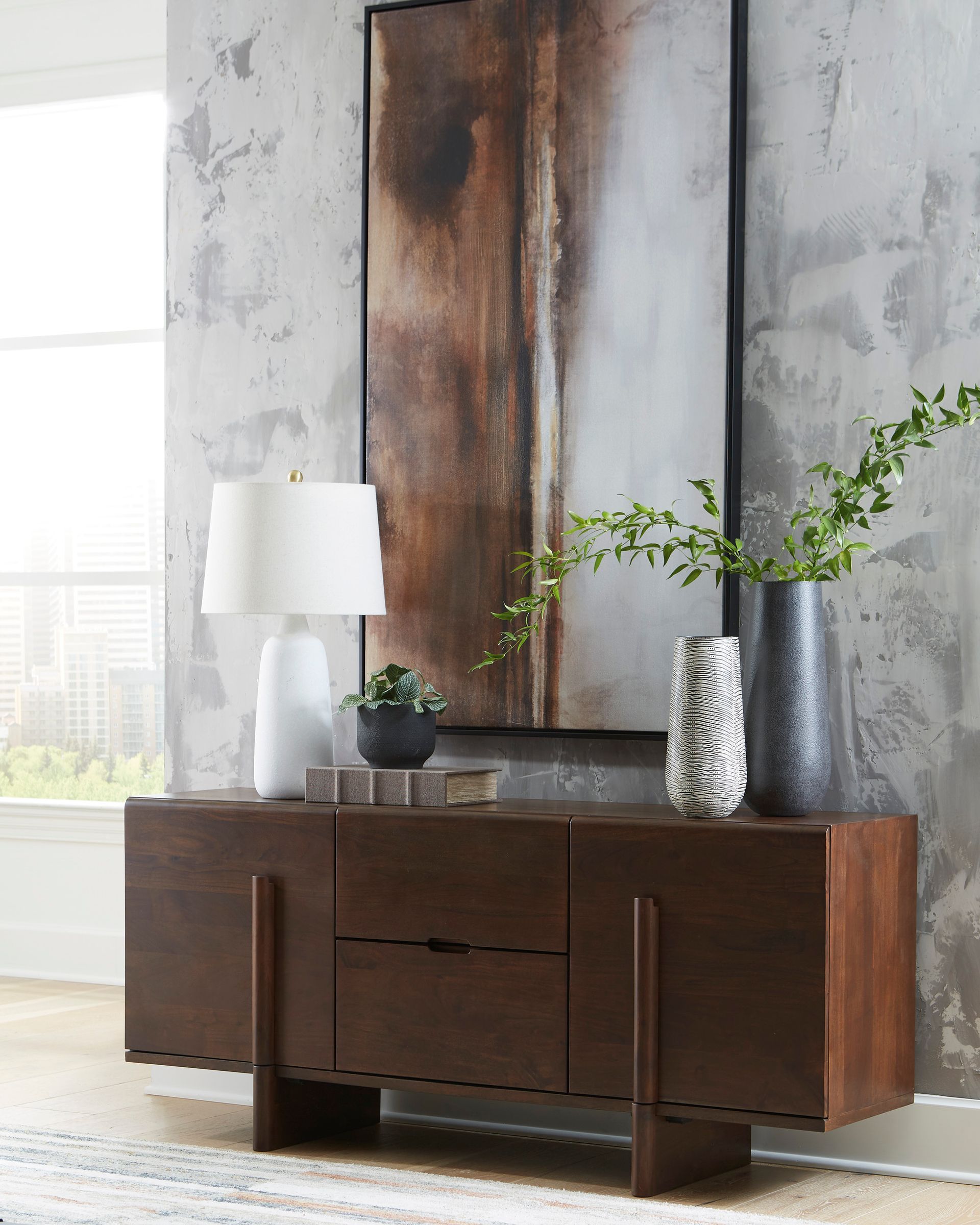 A wooden sideboard with artwork and decor against a textured gray wall, next to a window.