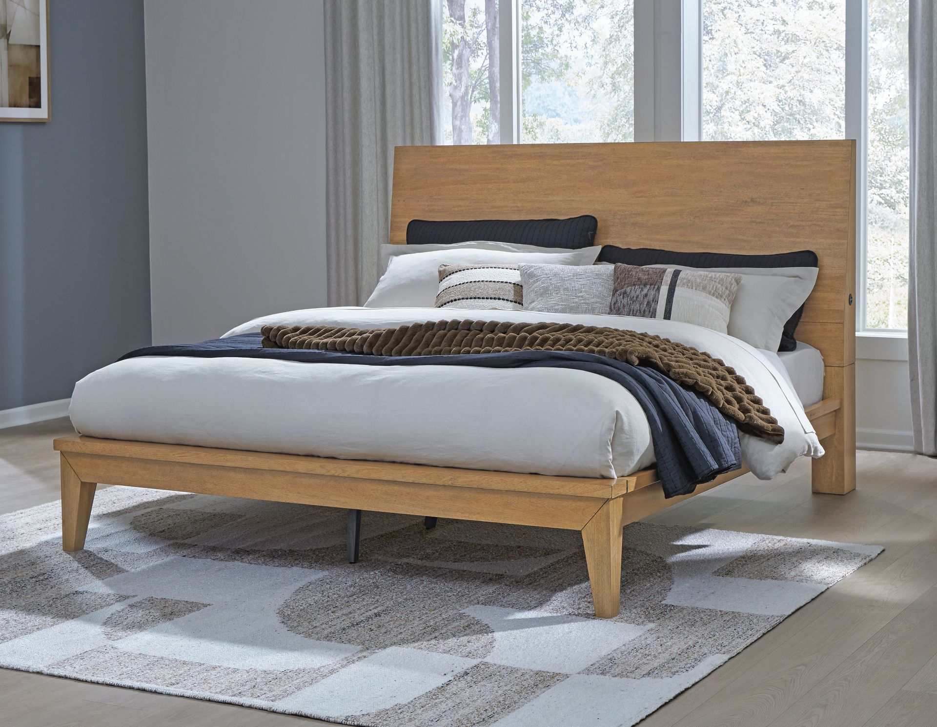 Wooden bed frame with large headboard, made up with white bedding and decorative pillows, on a patterned rug.