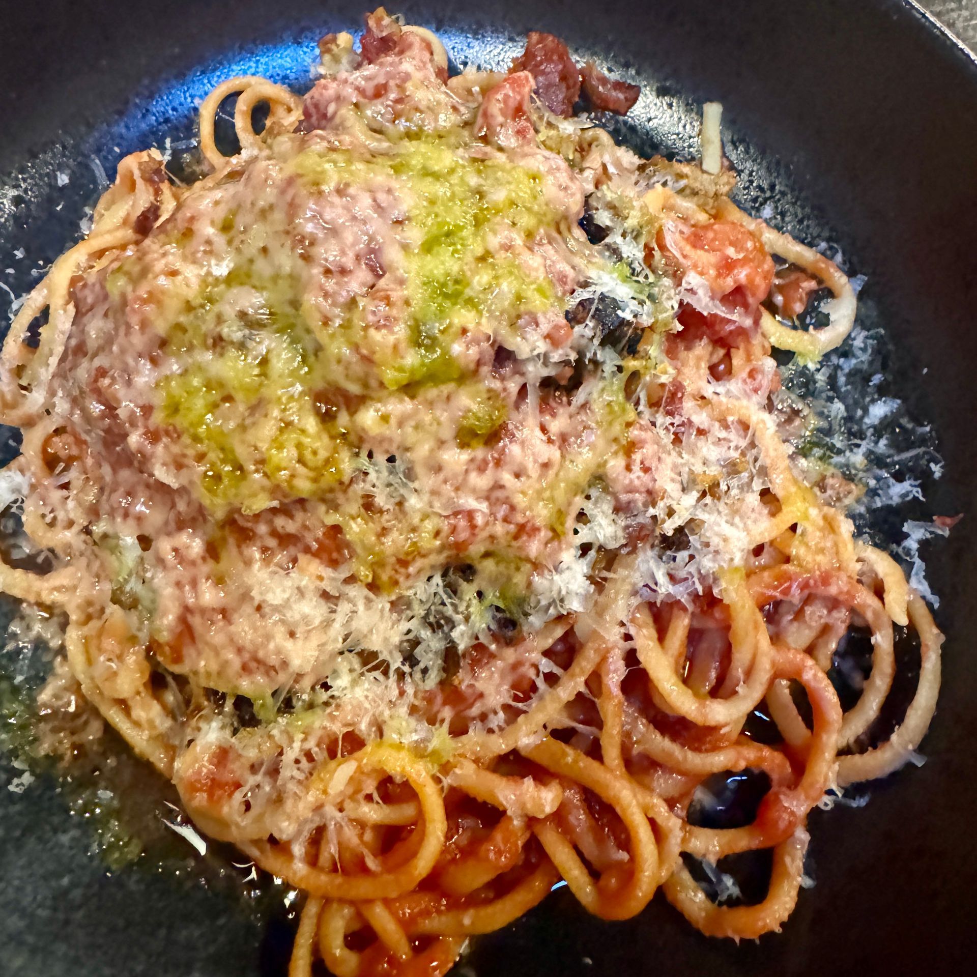 Spaghetti with tomato sauce, topped with pesto and grated cheese, on a dark plate.
