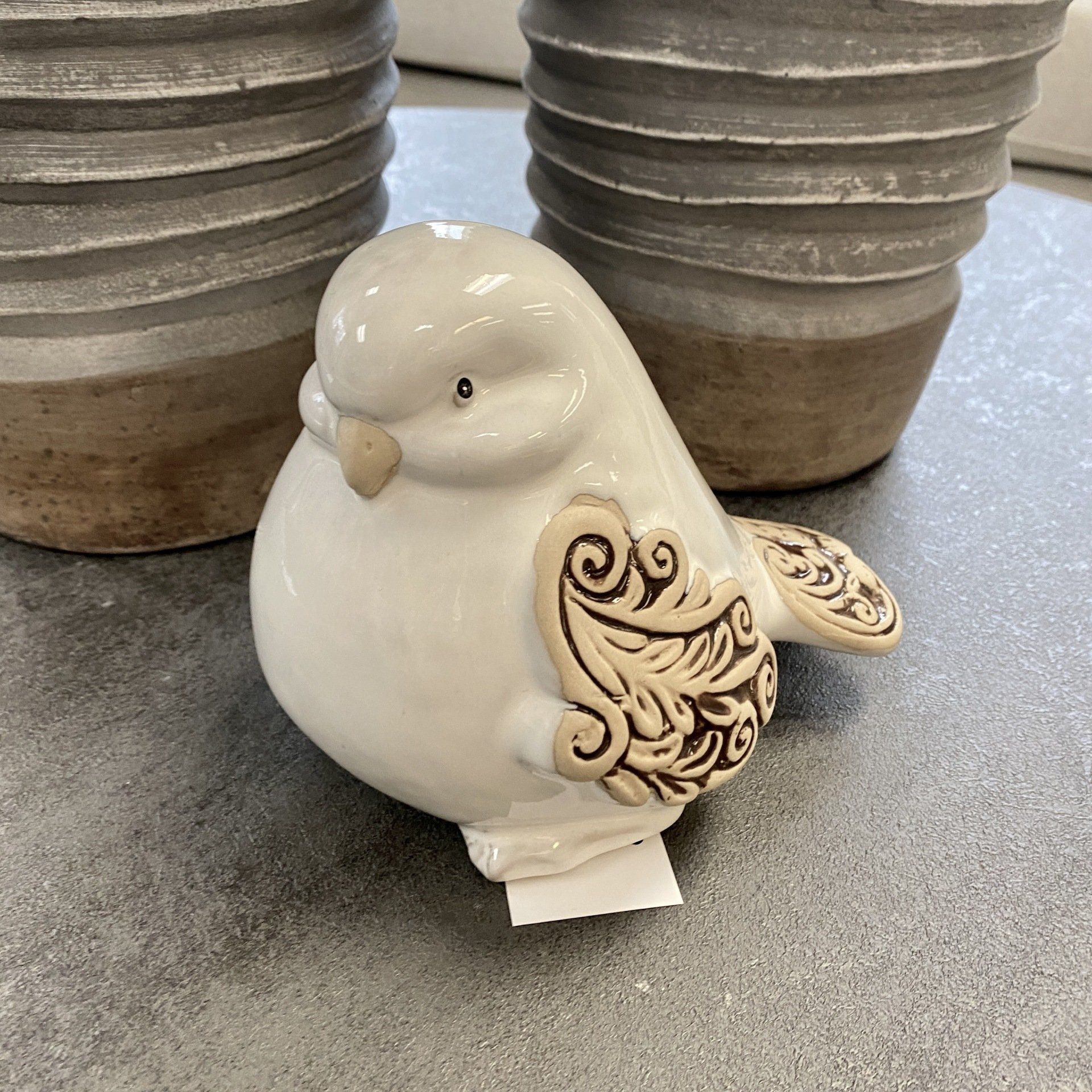 White ceramic bird figurine with brown wing detail, on a gray surface with two gray planters in background.