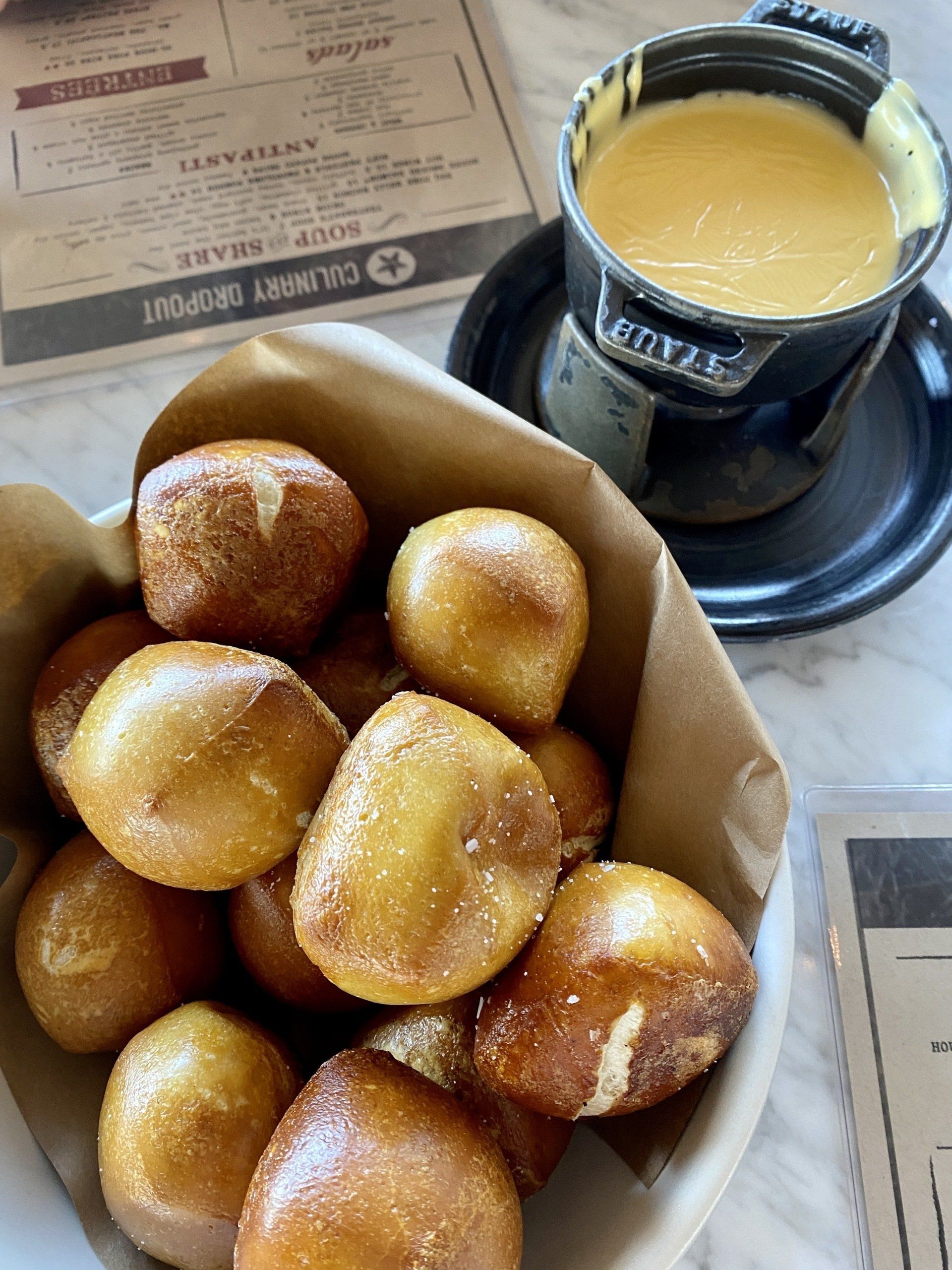 Pretzel bites in a paper-lined basket, served with cheese fondue in a black pot on a table.