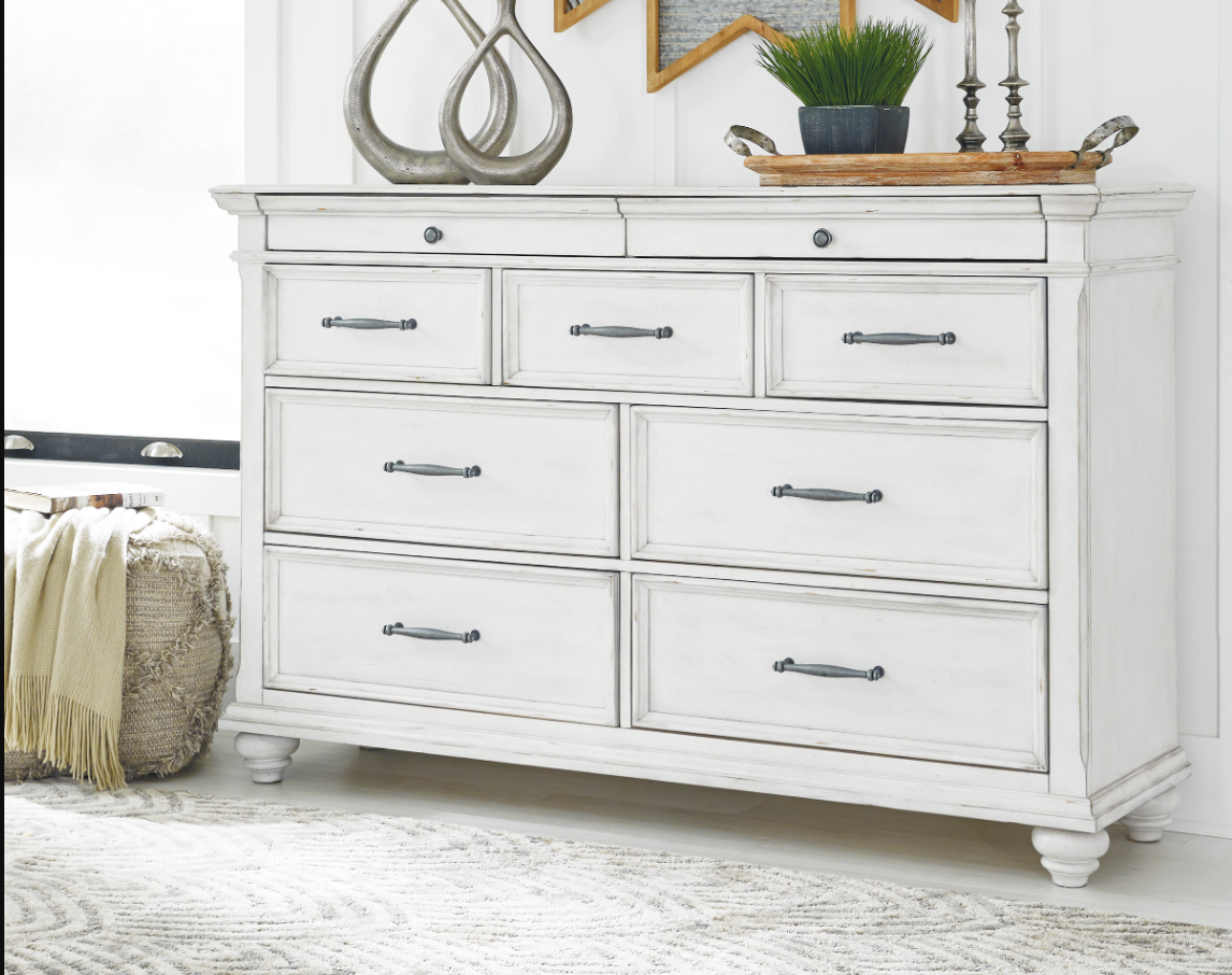 White distressed dresser with 9 drawers, holding decor, in a room with a rug and stool.