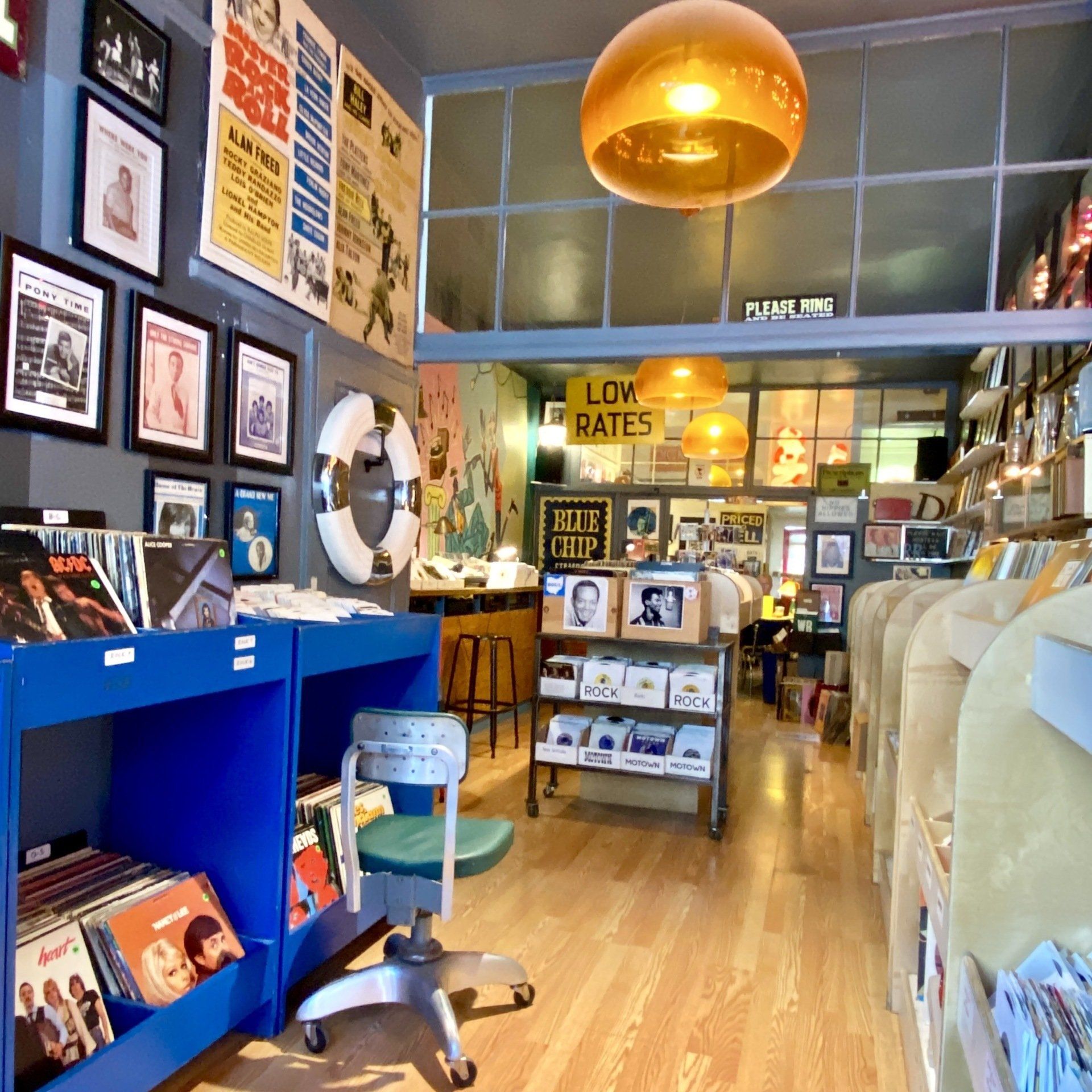 Inside a vintage record store: Blue shelves with albums, posters on the walls, and a hanging orange light.