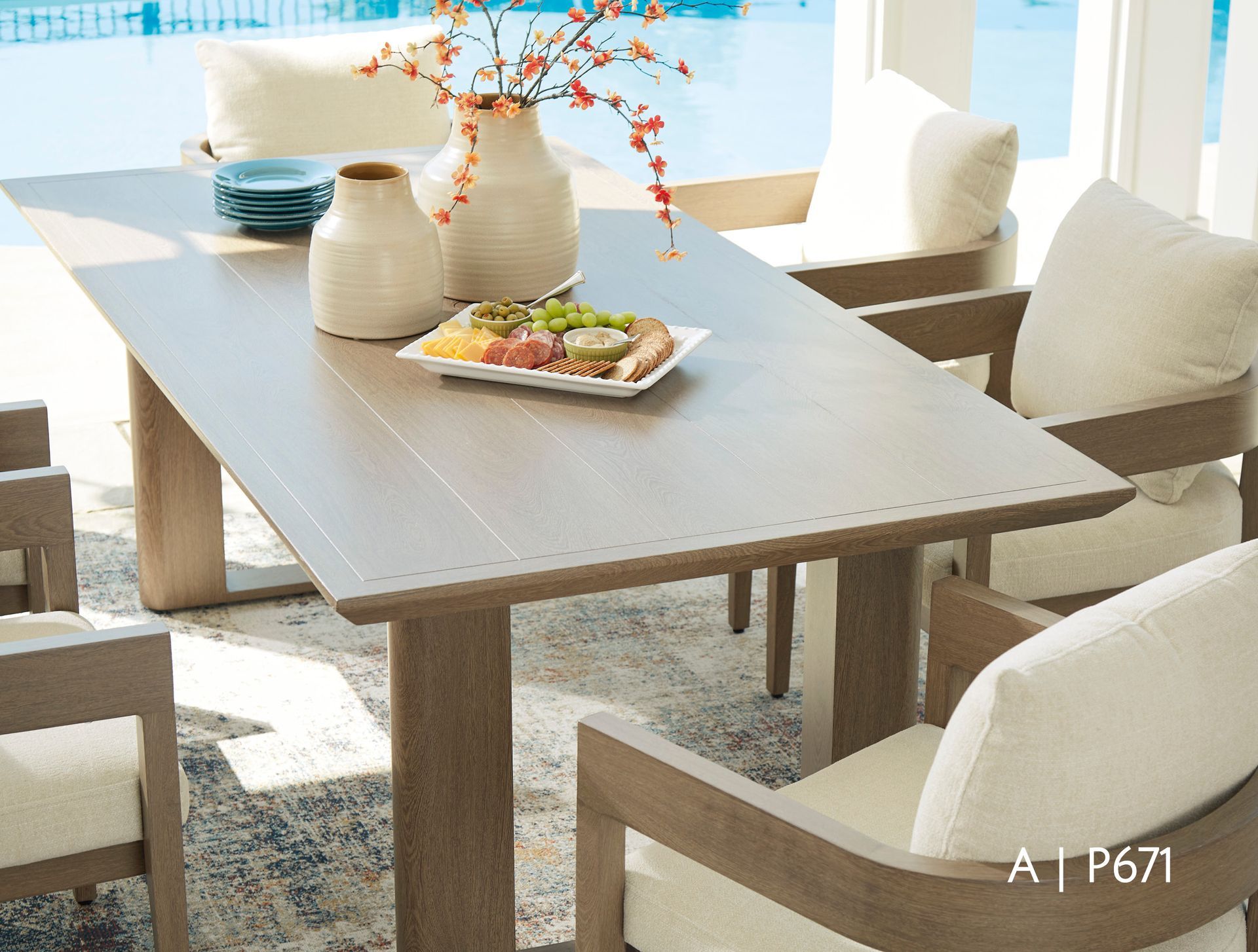 Wooden outdoor dining table with chairs, pillows, food, and vases.
