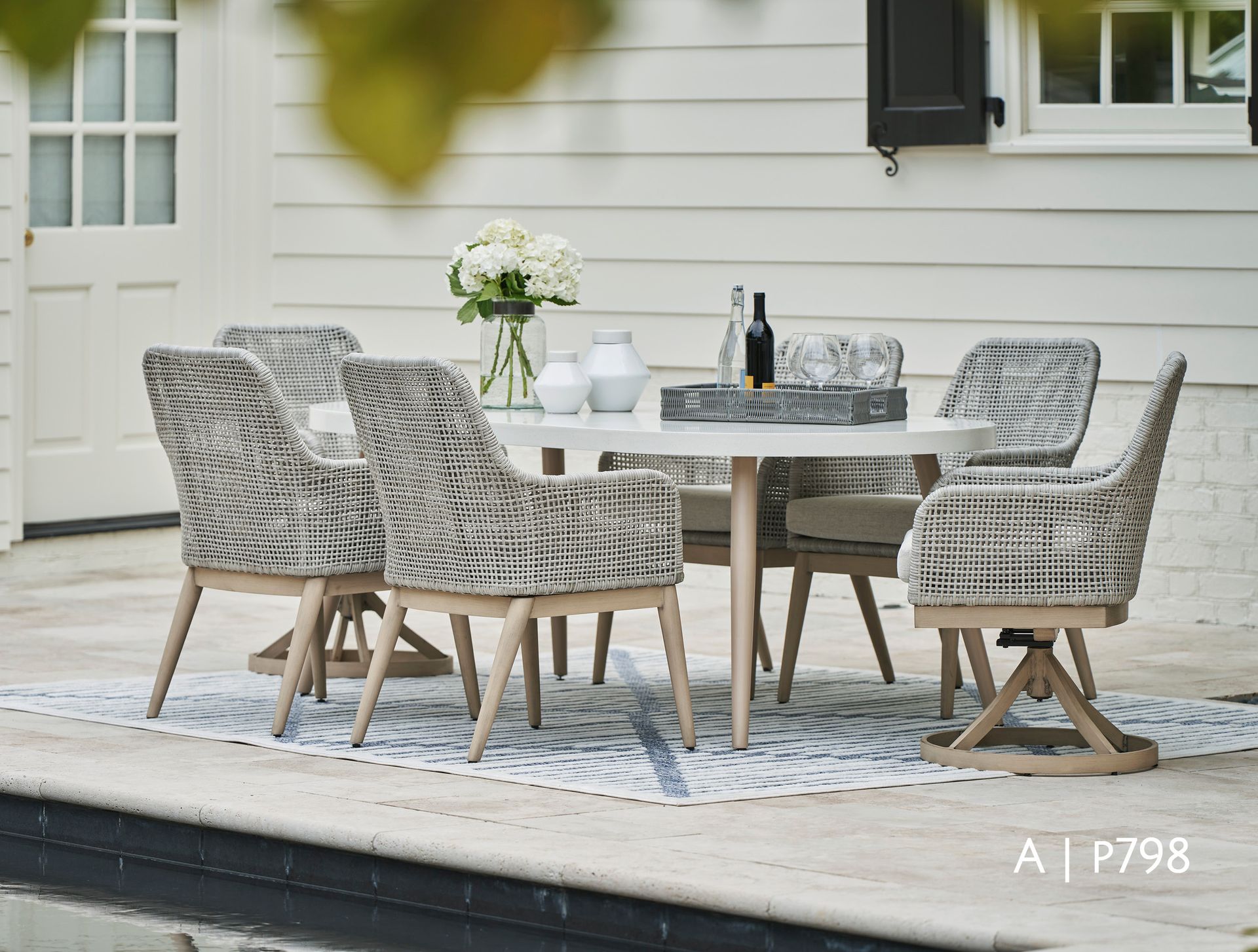Outdoor dining set: white table, woven chairs, rug, flowers, and wine on a patio.