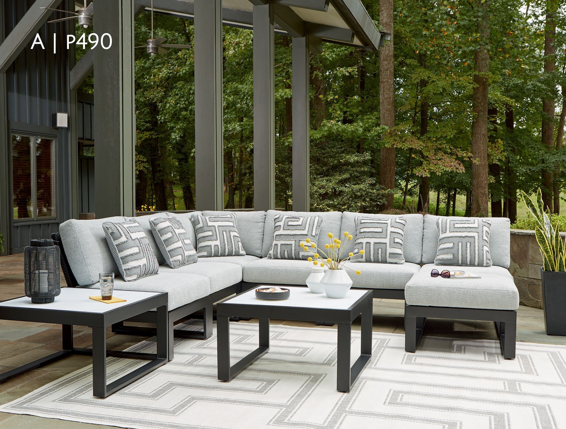 Outdoor sectional sofa with patterned pillows, tables, and a rug. Trees in the background.
