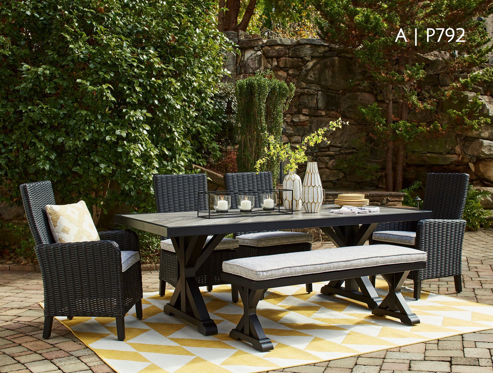Outdoor dining set: a black table, chairs, and bench on a patterned rug. Green foliage in the background.
