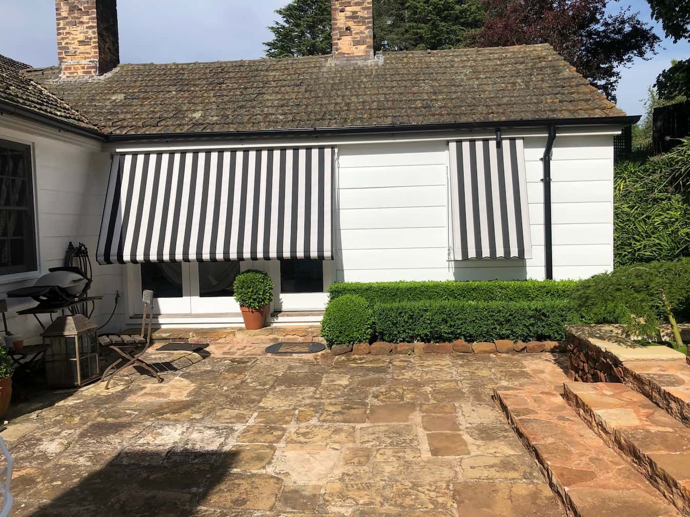White building with black and white striped awnings over windows. A stone patio and hedge are in front.