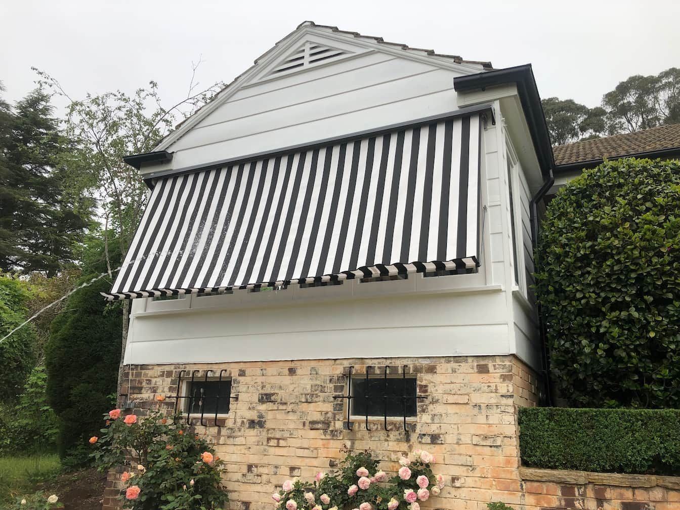 White house with black and white striped awning. Brick base, rose bushes, and greenery.
