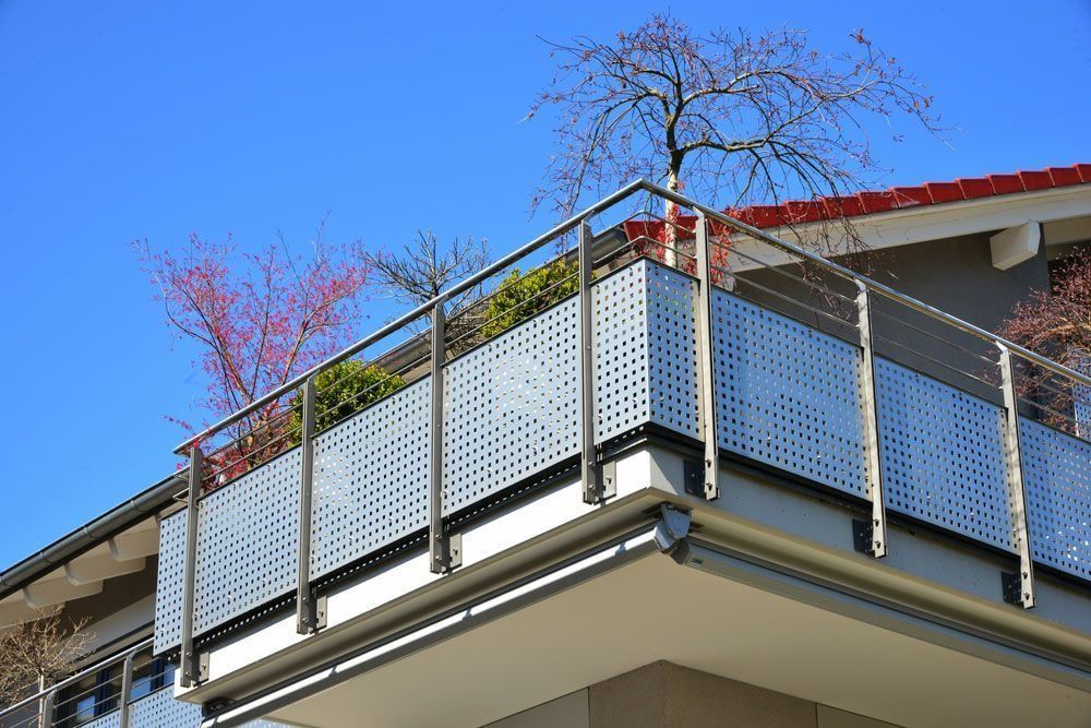 Balcony with perforated metal railing and small trees against a blue sky.