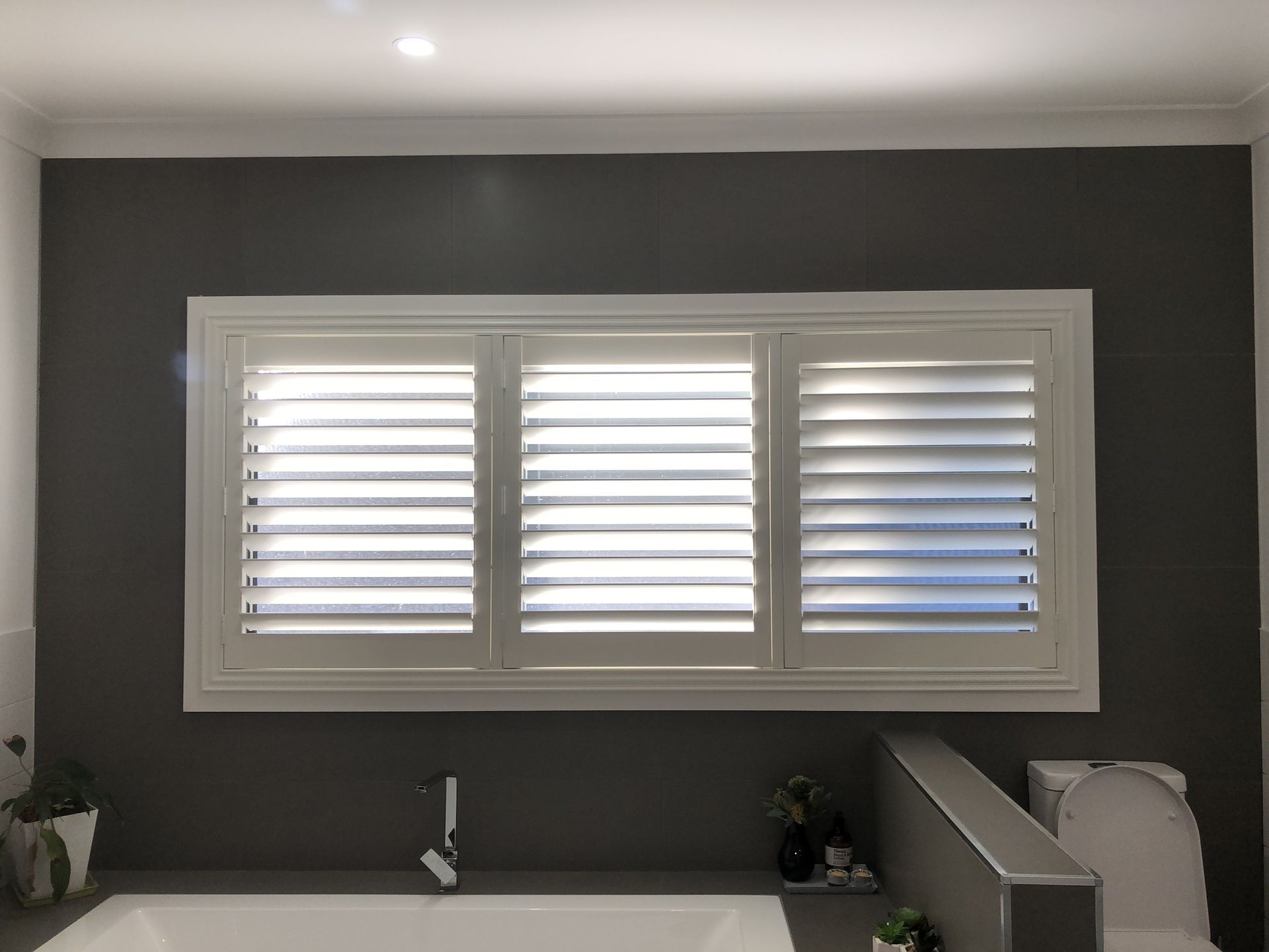 White shuttered windows over a gray bathroom wall. A bathtub is below.