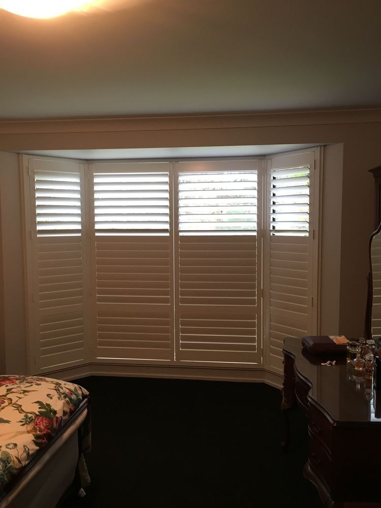 White shuttered window in a room with a bay window, with a bedroom interior visible.