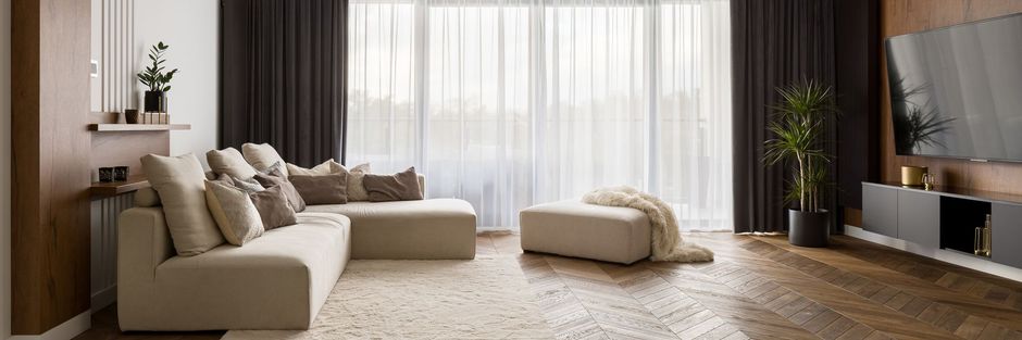 Living room with beige sectional sofa, ottoman, floor-to-ceiling windows, dark curtains, and wood flooring.