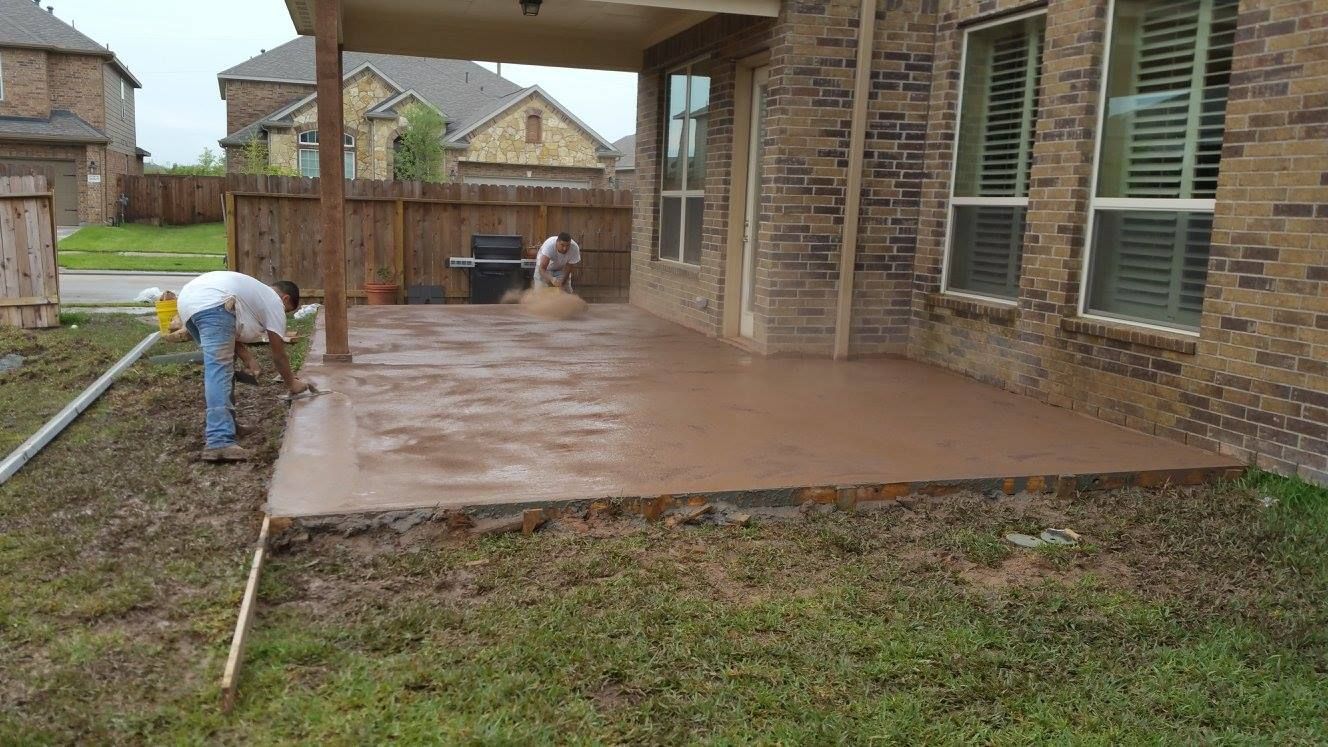 Workers pouring and smoothing fresh concrete patio next to a brick house on a cloudy day.