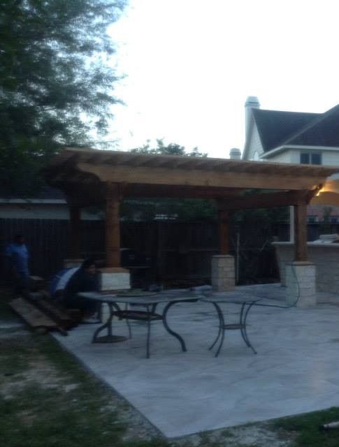 Pergola with dining table and chairs on a concrete patio. House in the background.