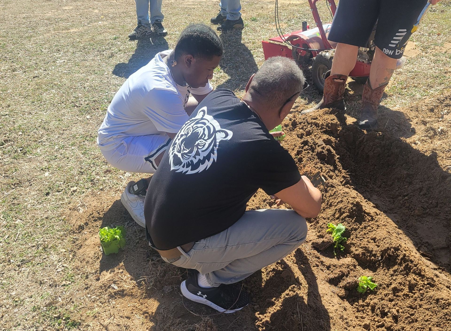 A man in a black shirt with a tiger on it is kneeling in the dirt.