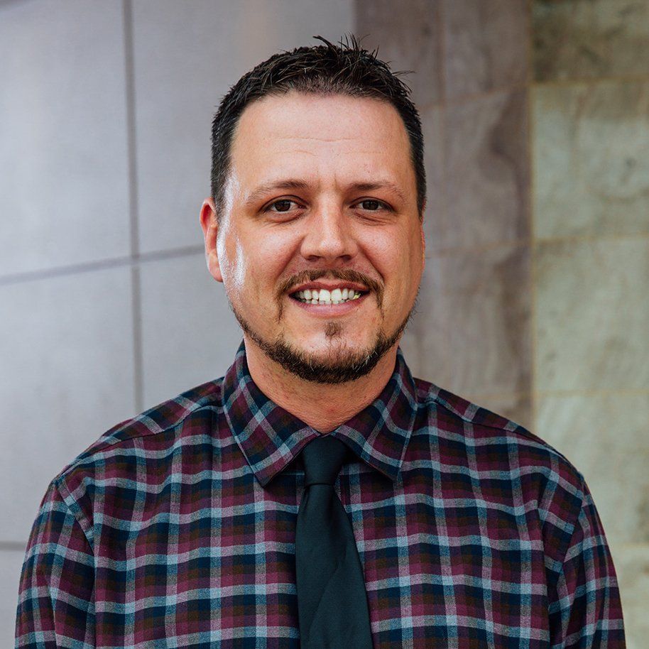 A man with a beard wearing a plaid shirt and tie is smiling for the camera.