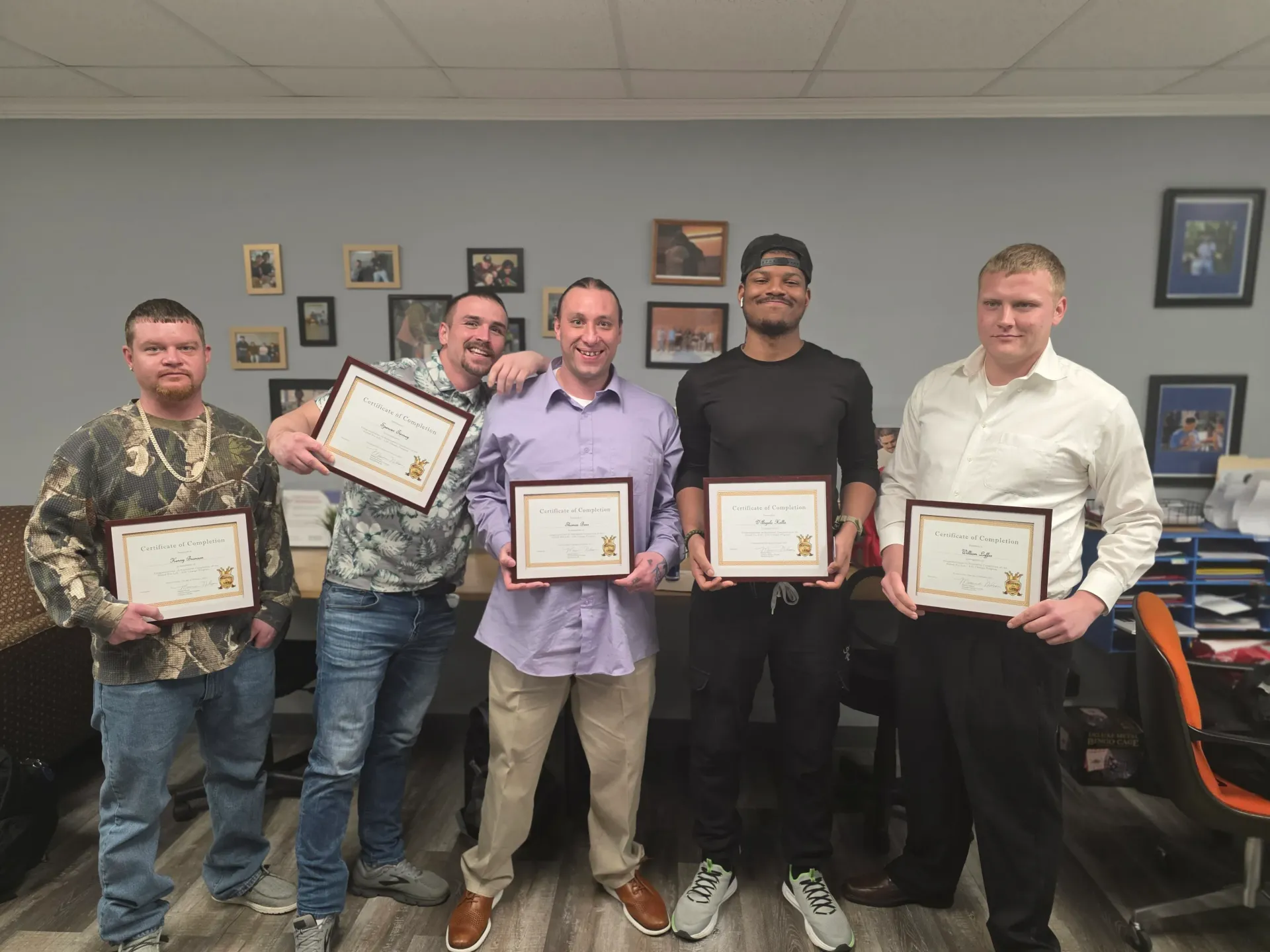 Five people holding framed certificates stand smiling in an office setting.