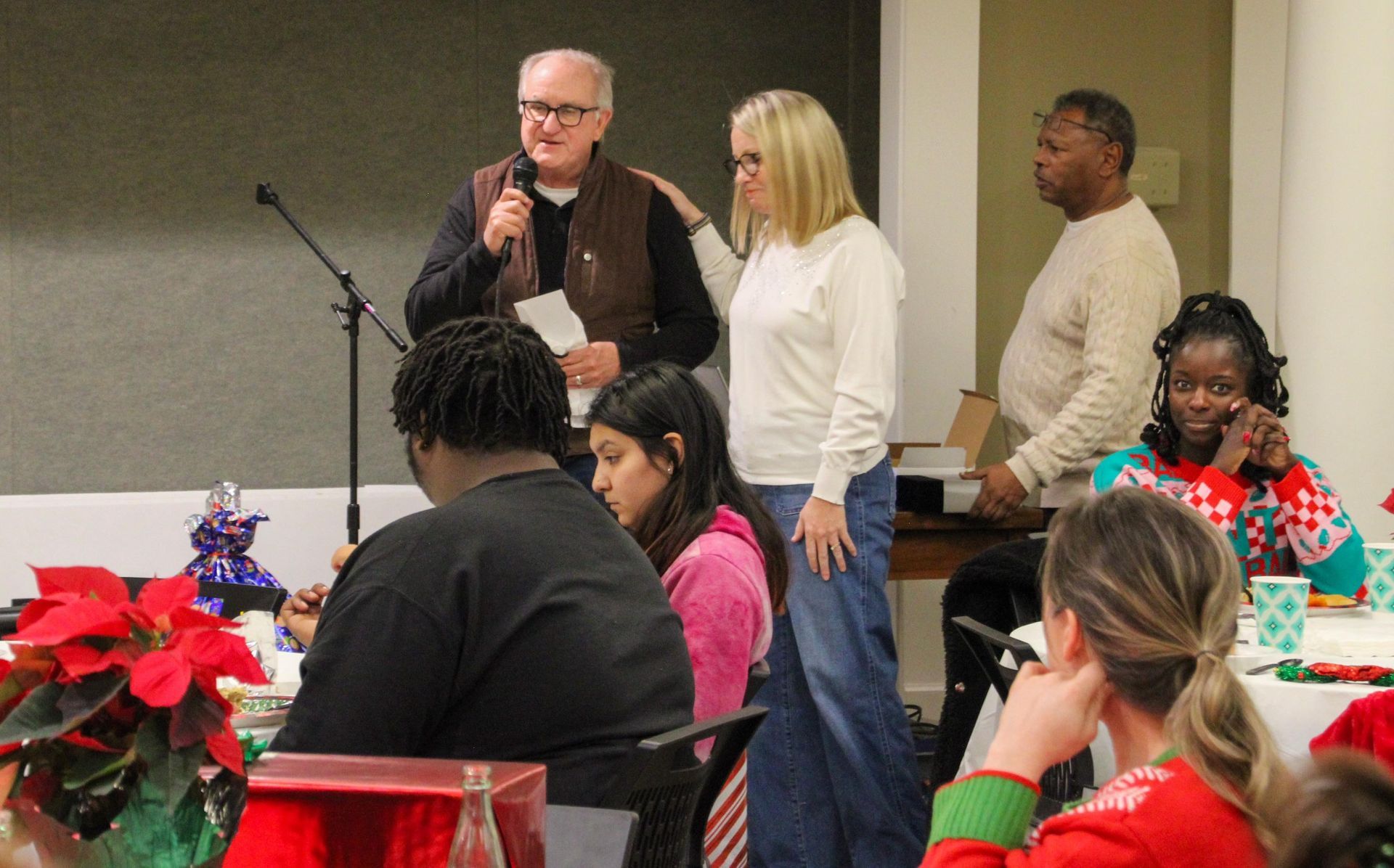 A man is standing in front of a microphone talking to a group of people during a holiday party.