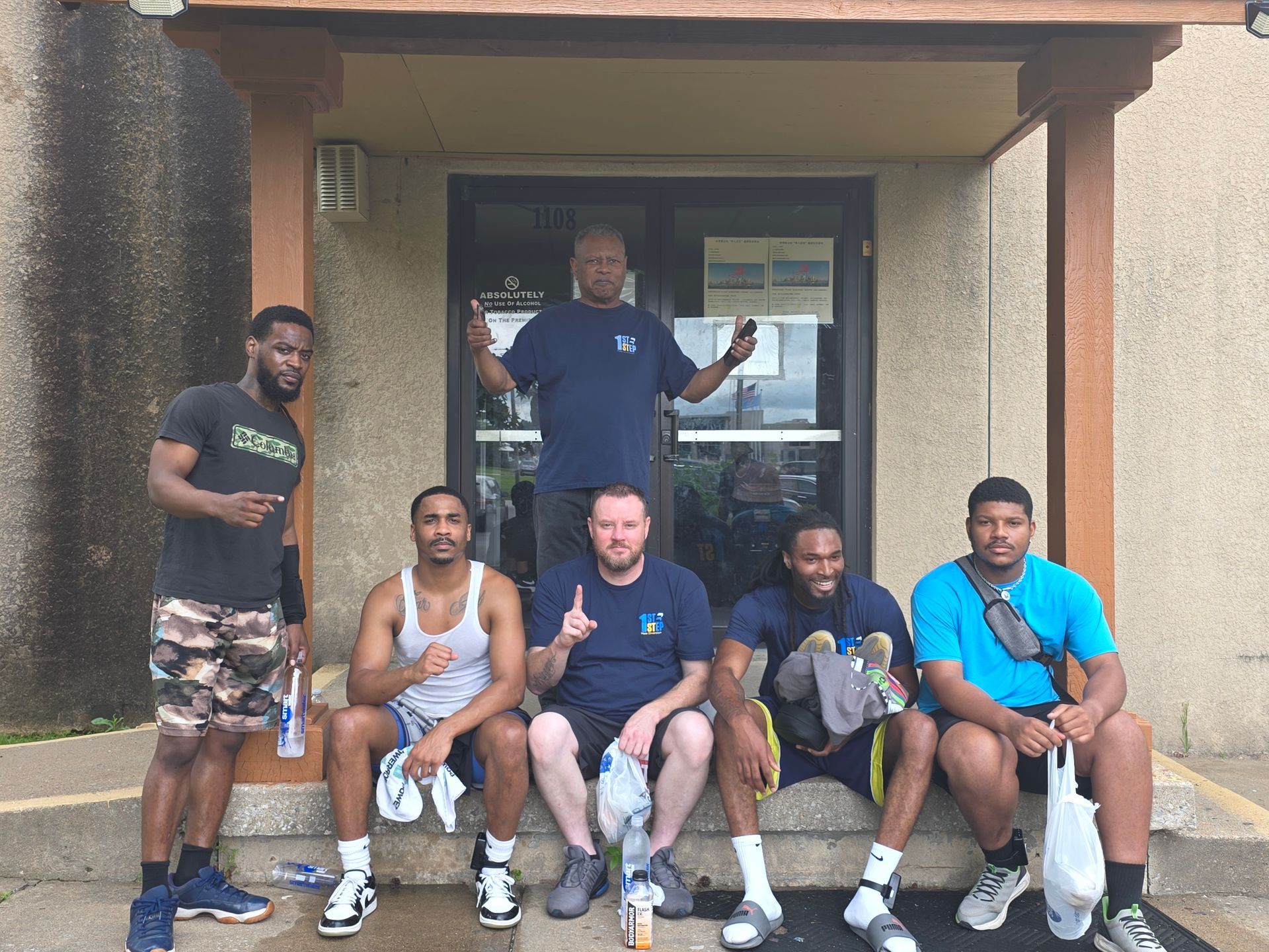 Six men posing on a front porch entrance, one standing behind five seated on the steps.