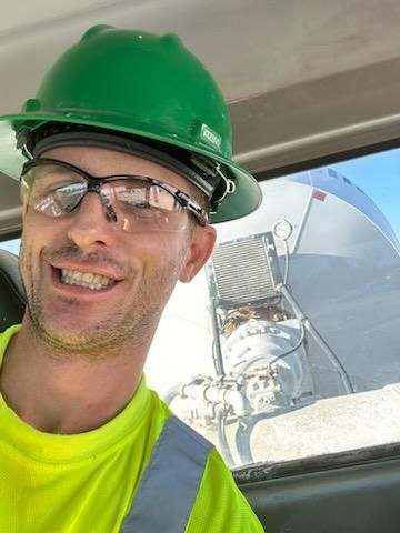 A man wearing a green hard hat and safety glasses is smiling.