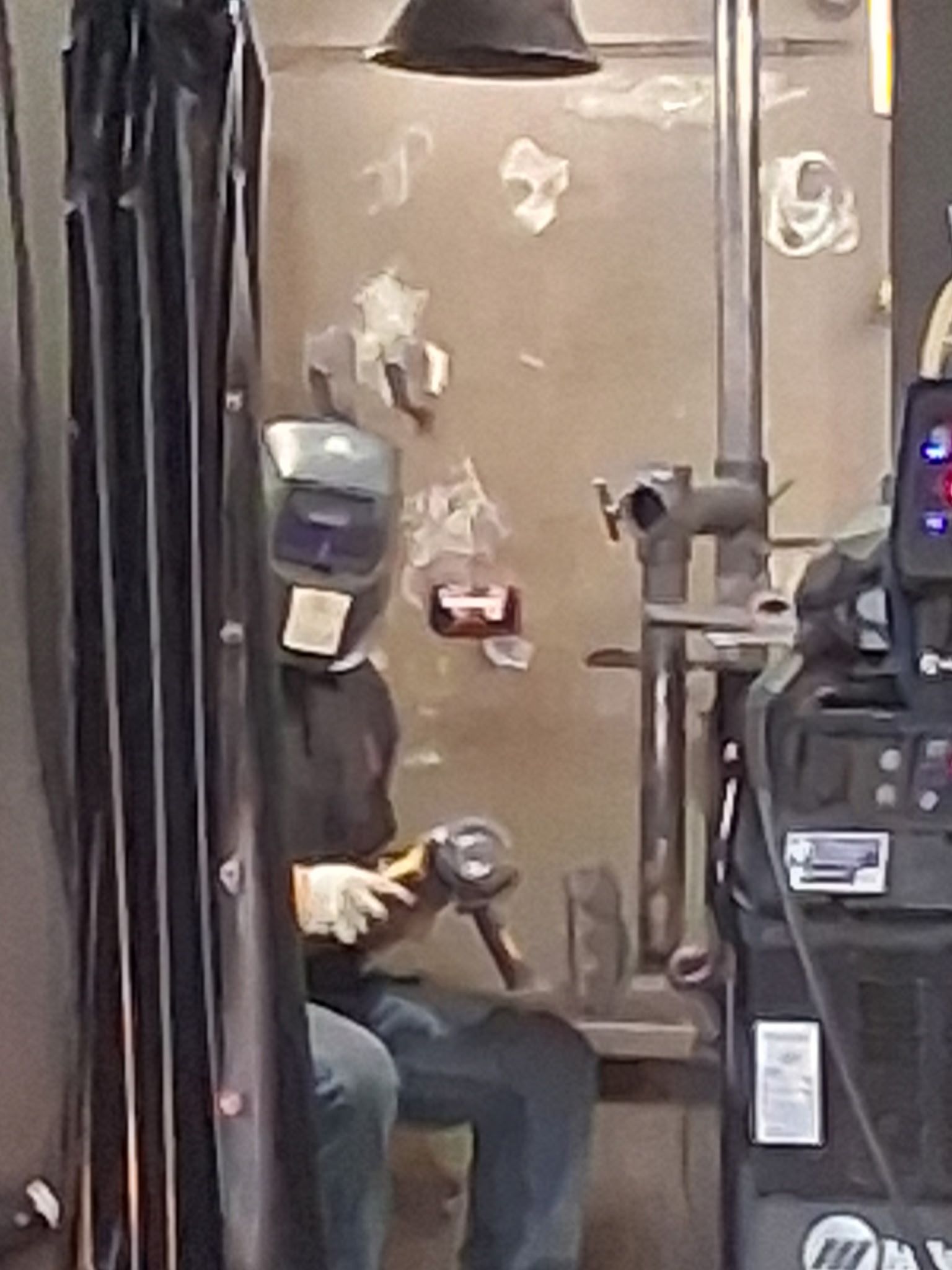 Person in a welding helmet sits indoors, holding a tool beside a wall with peeling paint.