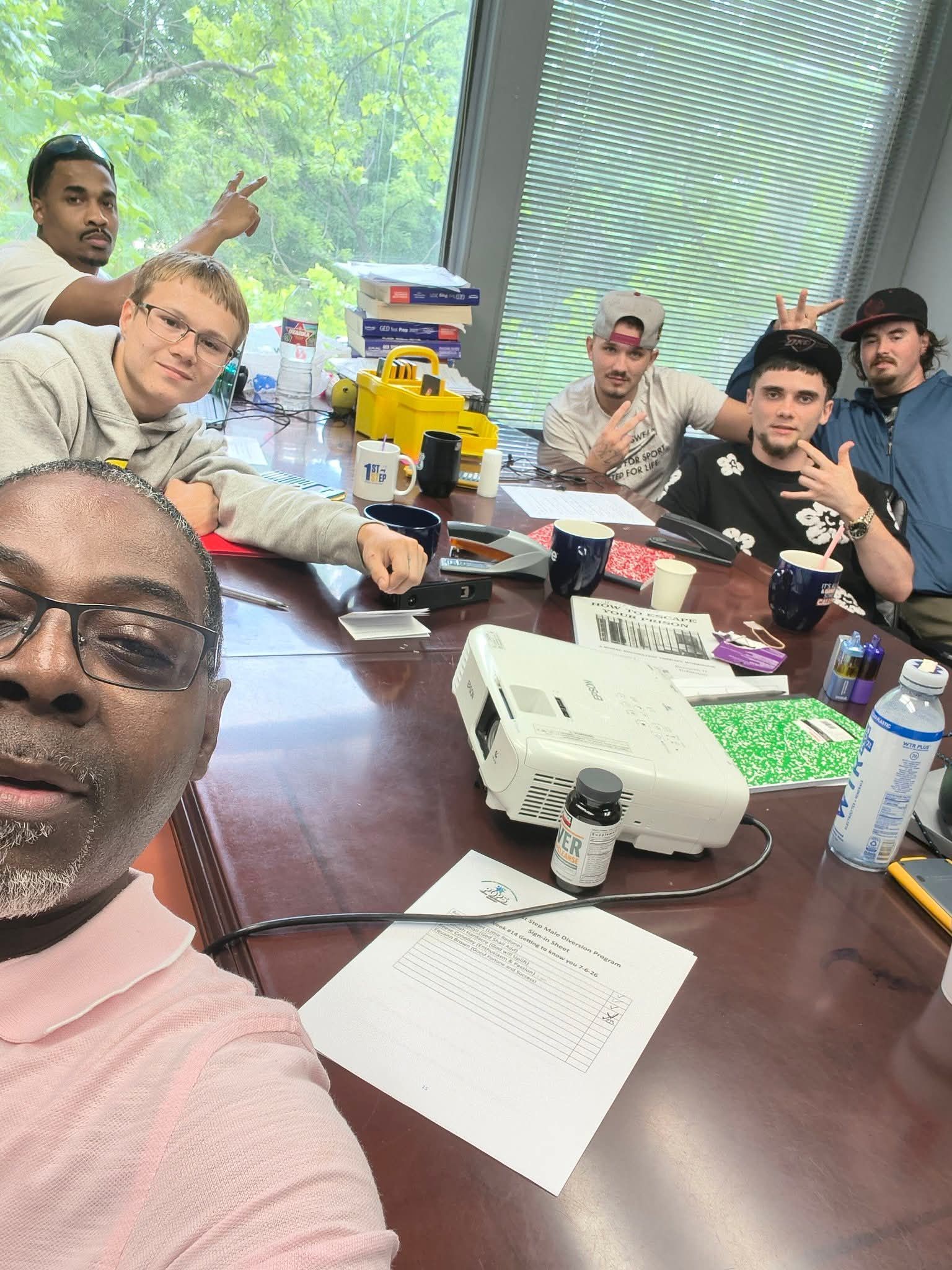 Group selfie in a meeting room around a conference table with laptops, papers, and a projector.