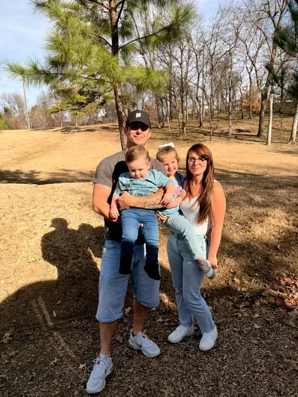 Family of four posing outdoors in a park, holding two children under a bright sky.