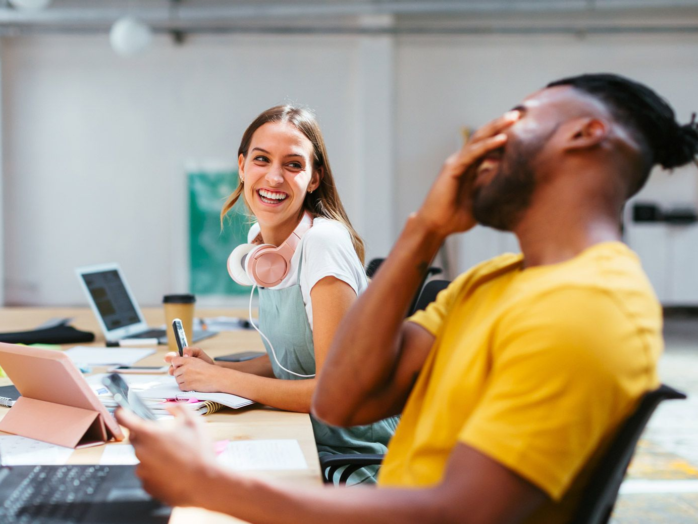 Two coworkers smiling in a bright office, one in a yellow shirt covering his face with laughter