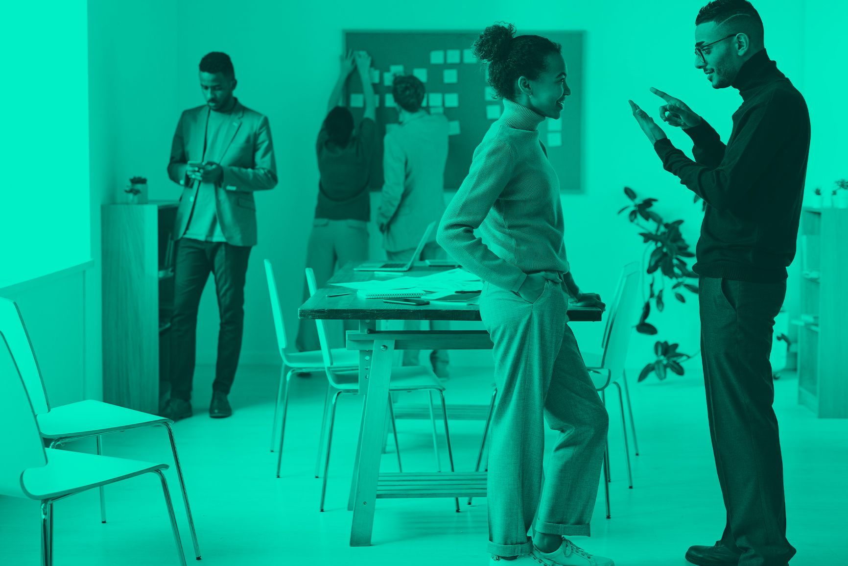 People conversing in a modern office, with one standing and talking to another near a table and chairs.