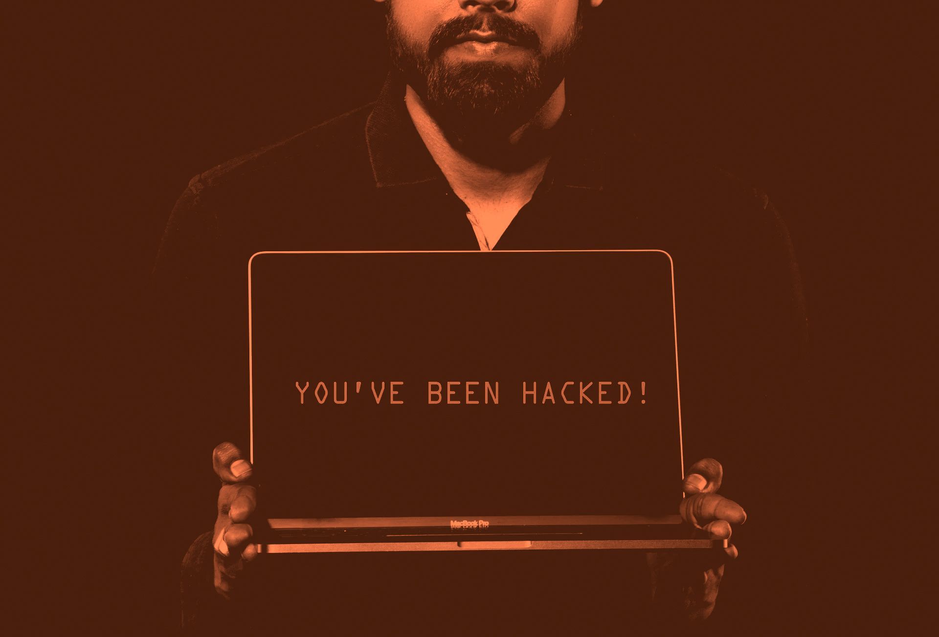 Person holding an open laptop with “YOU’VE BEEN HACKED!” on a dark red background