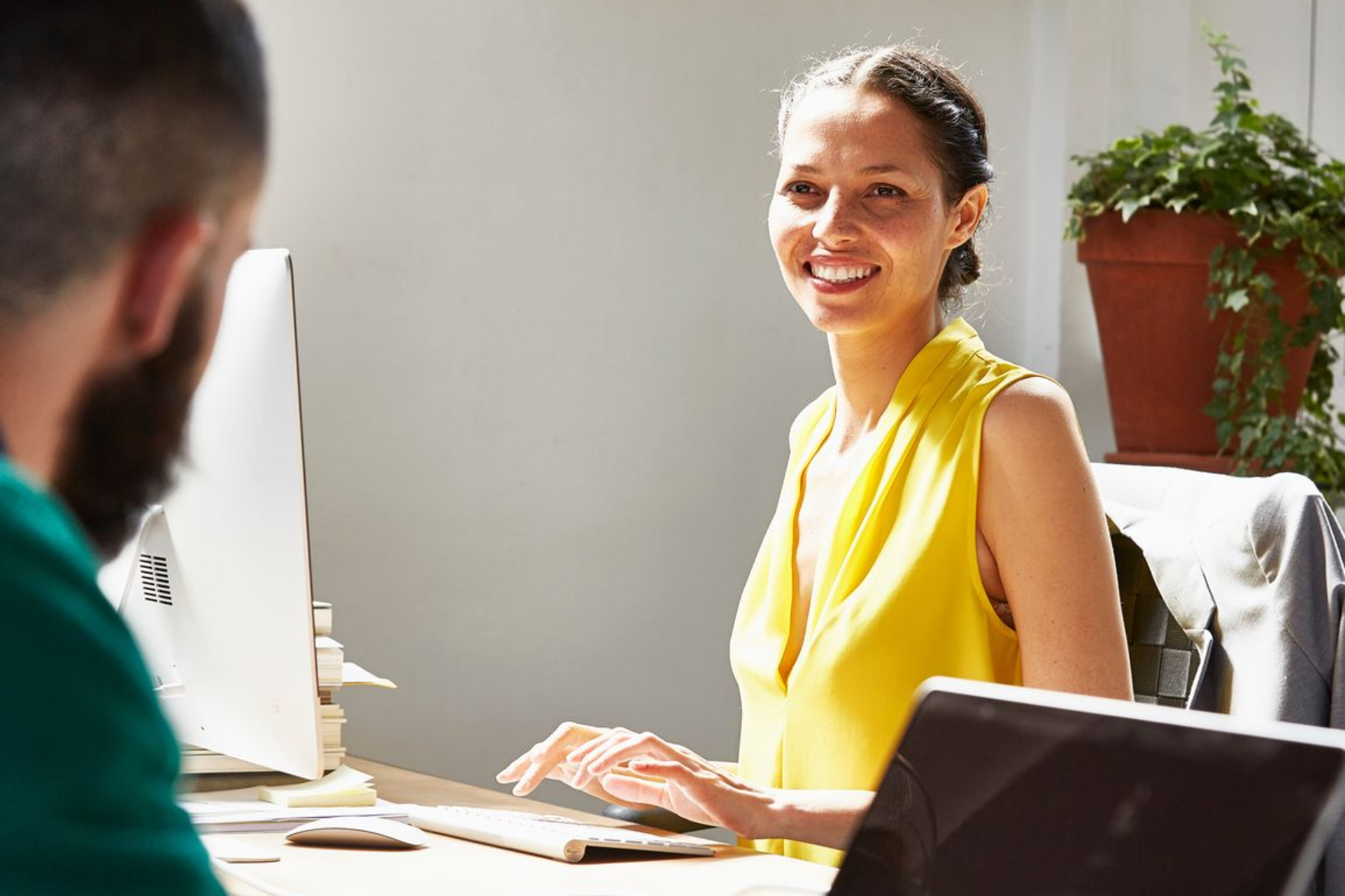Two people meeting at a sunlit office table, with a laptop, papers, and a smiling woman in a yellow top