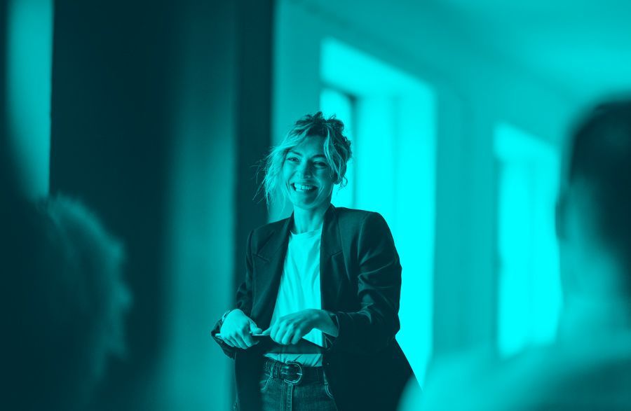 Person standing indoors in teal lighting, speaking while holding a remote, with blurred audience in foreground