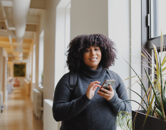 Person in a hallway looking at a phone, wearing a dark sweater beside a window and plant.