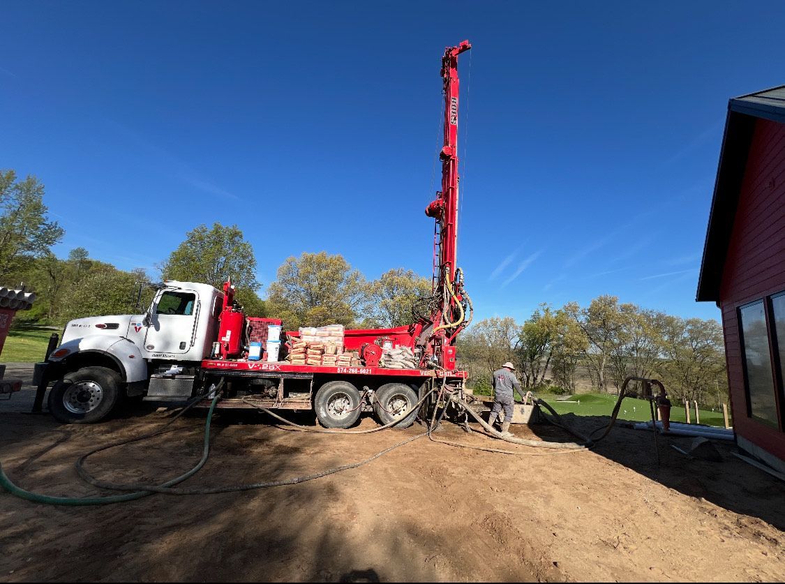 Red and white drilling rig on brown dirt, drilling a well next to a red building under a blue sky.