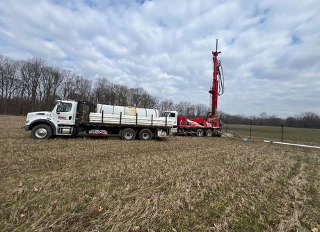 Truck and drilling rig in a field, preparing to drill. Red, white and green in a cloudy sky.