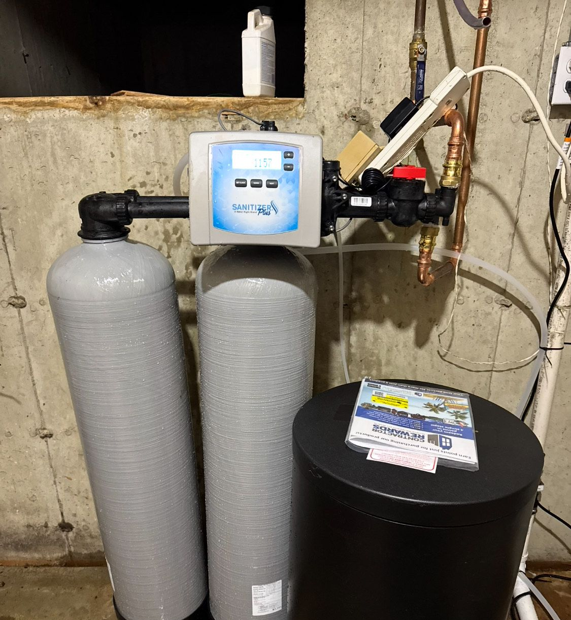 Water softener system in a basement. Gray tanks, digital control panel, black brine tank, and copper pipes.