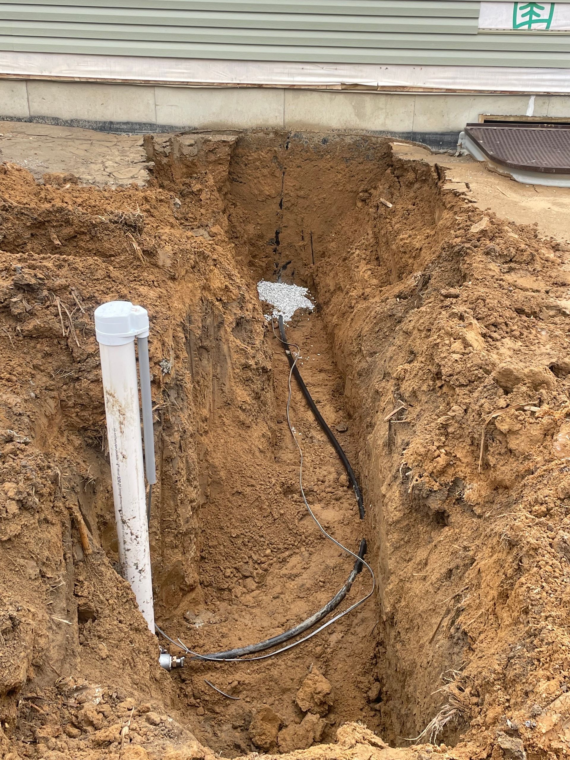 Hole in dirt revealing black cable and white pipe near a building.