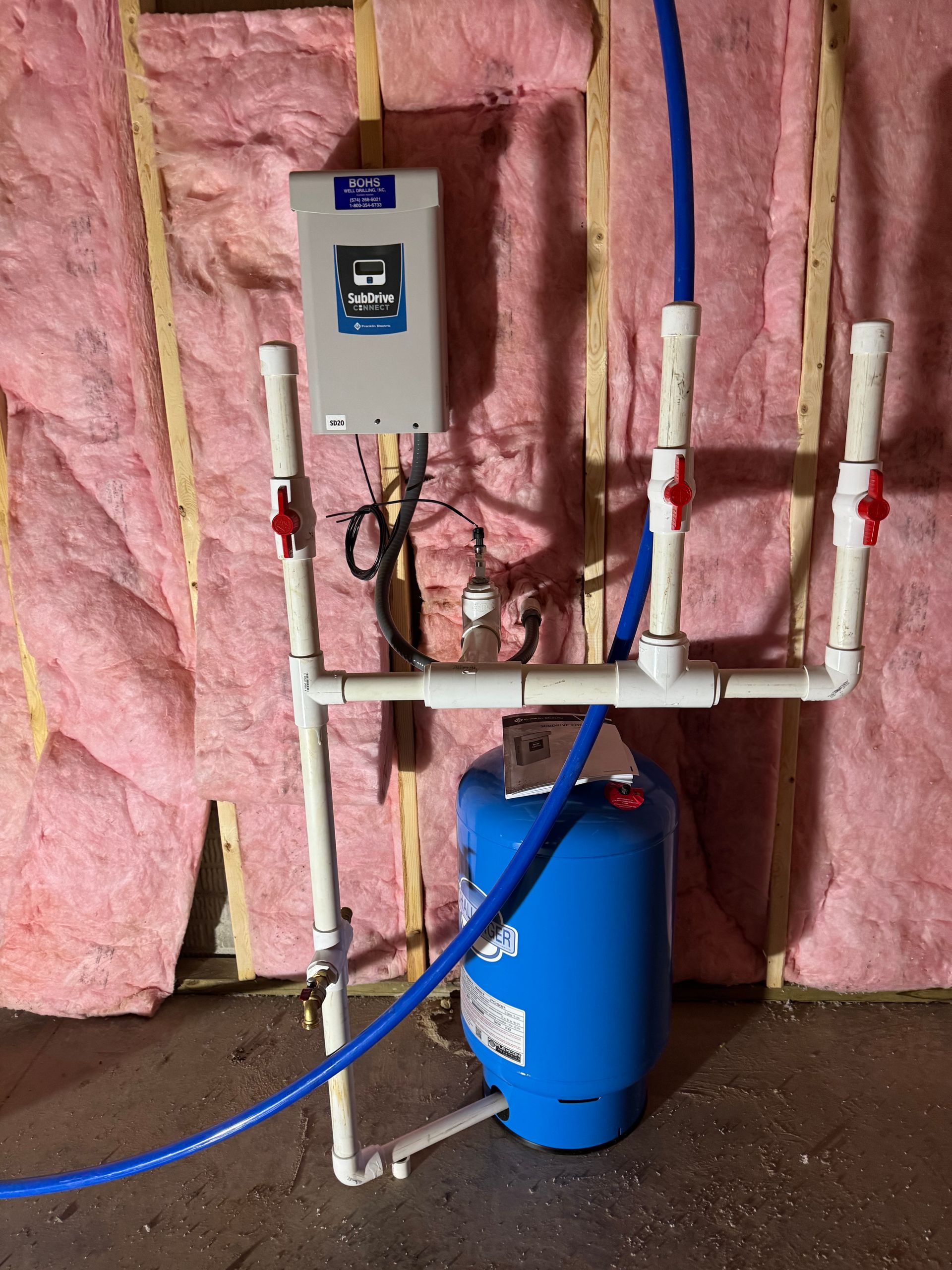 Blue water tank with white PVC pipes, electronic box, and pink insulation on a wall.