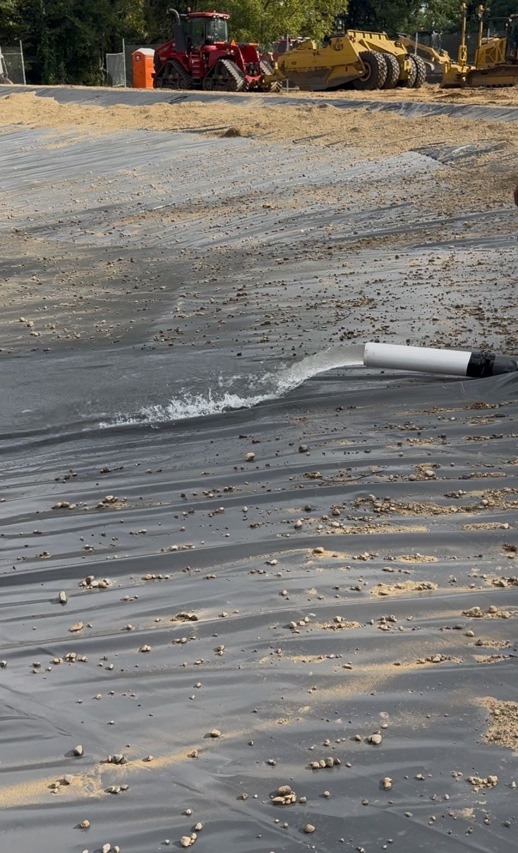Water spraying from a white pipe onto a black tarp at a construction site; heavy machinery in background.