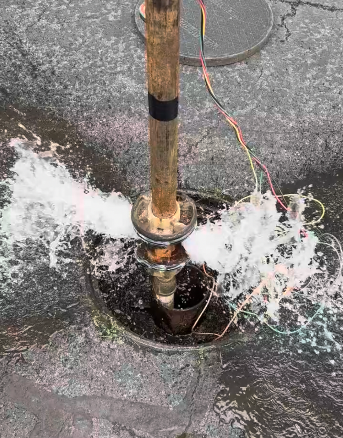 Water spurts from a well casing, surrounding a wooden pole with wires, on gray pavement.