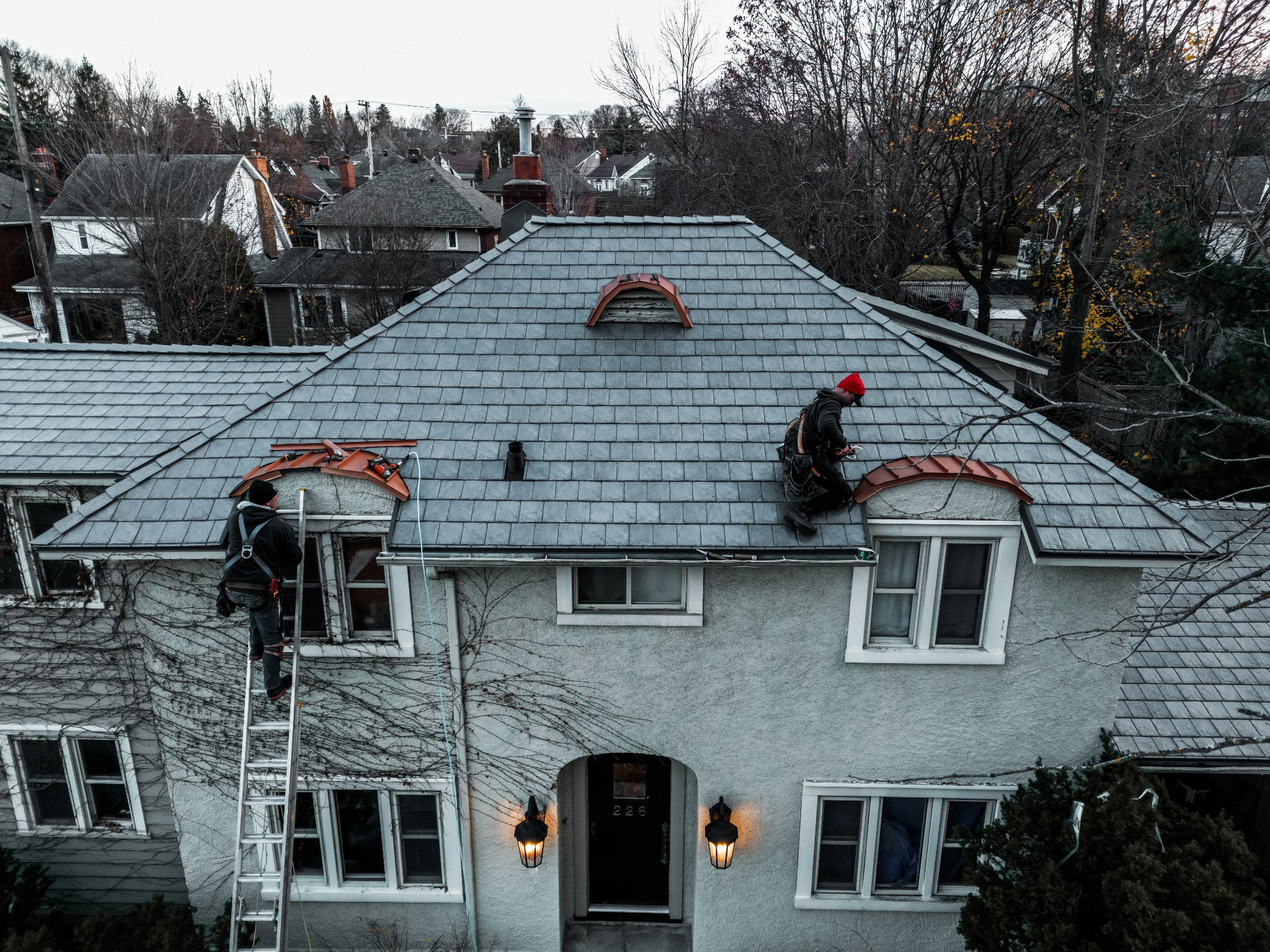 Two workers on a gray shingled roof, one in a red hat, on a house with dormer windows and lit entry lanterns.