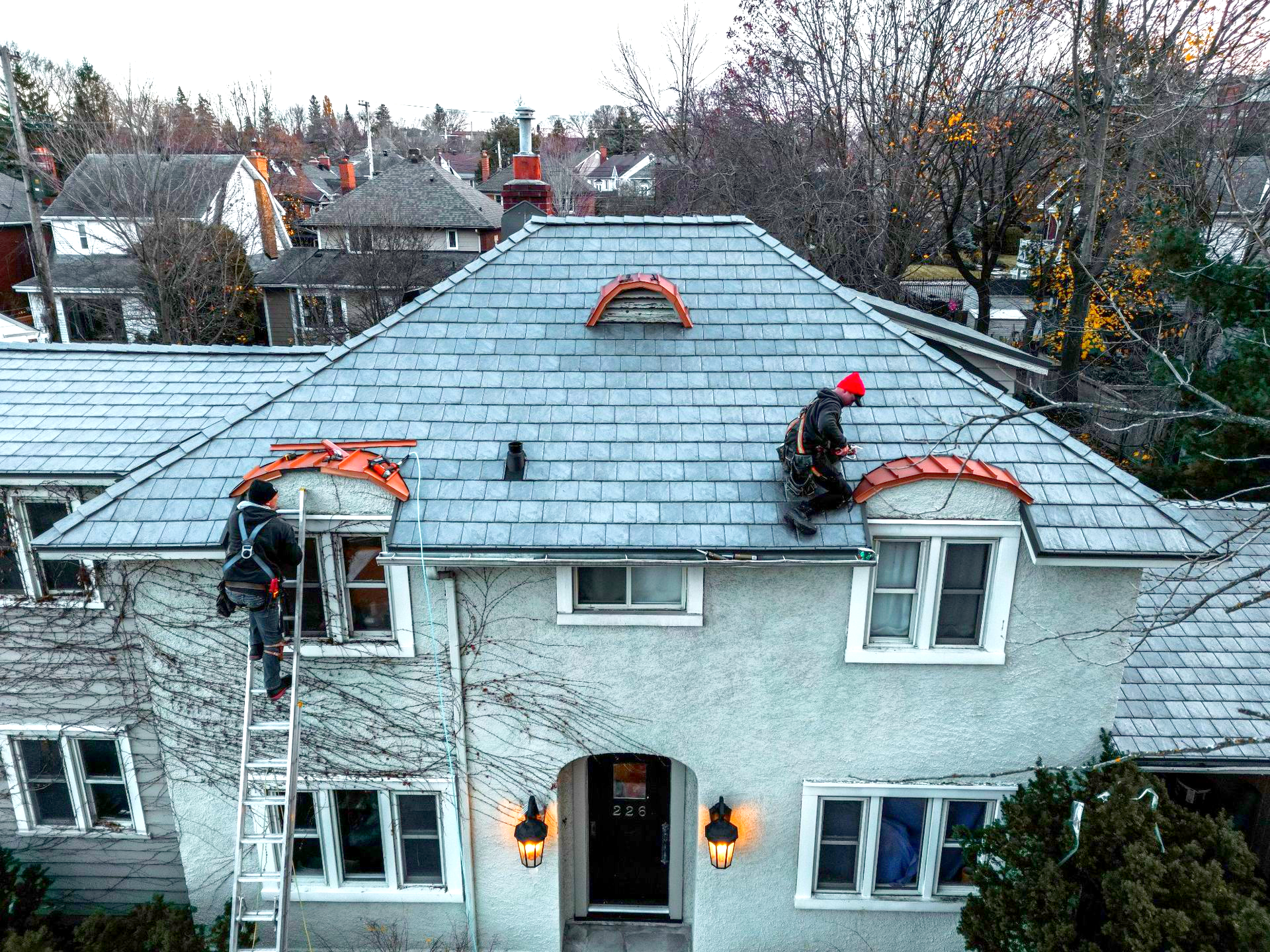 Workers on a snowy roof repairing a house, with ladders and tools visible.