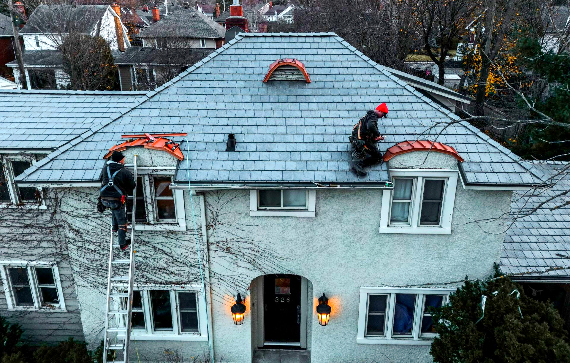Workers on a gray house roof installing shingles, with red safety helmets and ladders visible