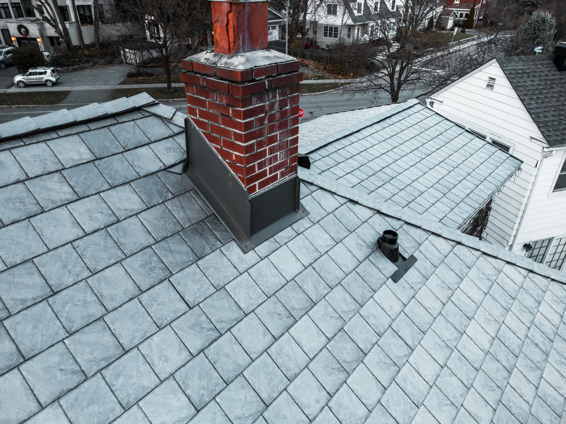 Snow-covered rooftop with a brick chimney and gray shingled roofs in the background