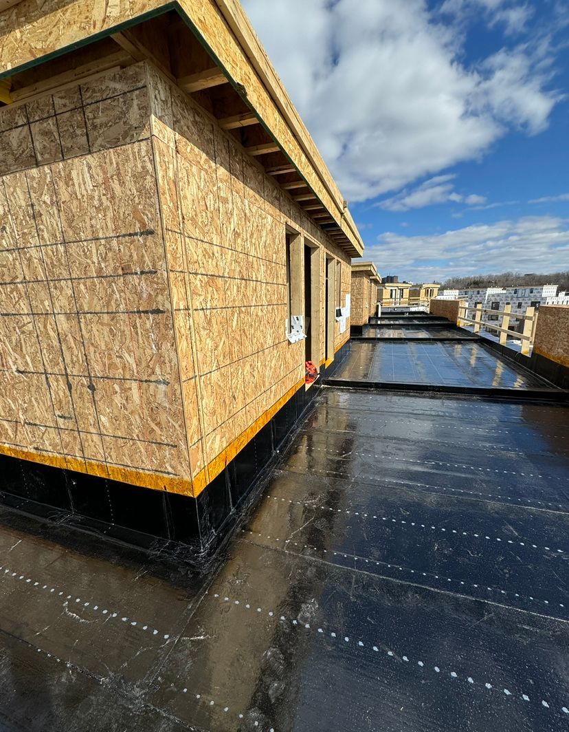 Rooftop under construction with plywood walls and black waterproofing, reflecting sunlight under a blue sky