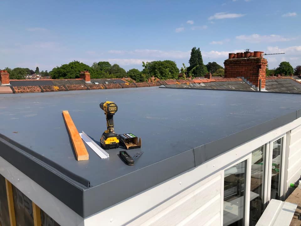 Roof with gray membrane, tools, and tape measure beside a low white building under a blue sky.
