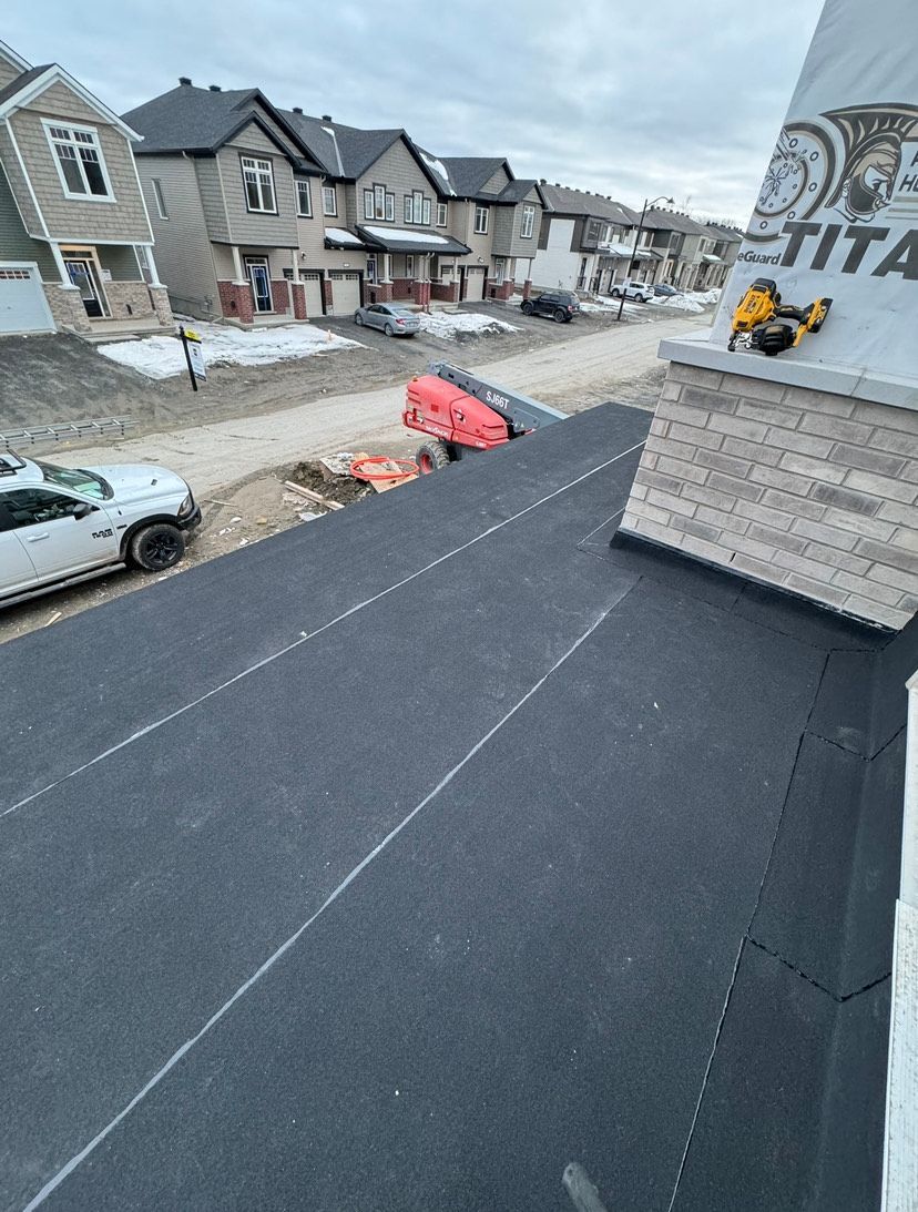 Flat black roof under construction beside snowy suburban homes and parked cars.