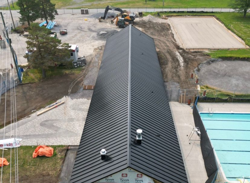 Aerial view of a building roof with dark shingles, surrounded by construction equipment and dirt work.